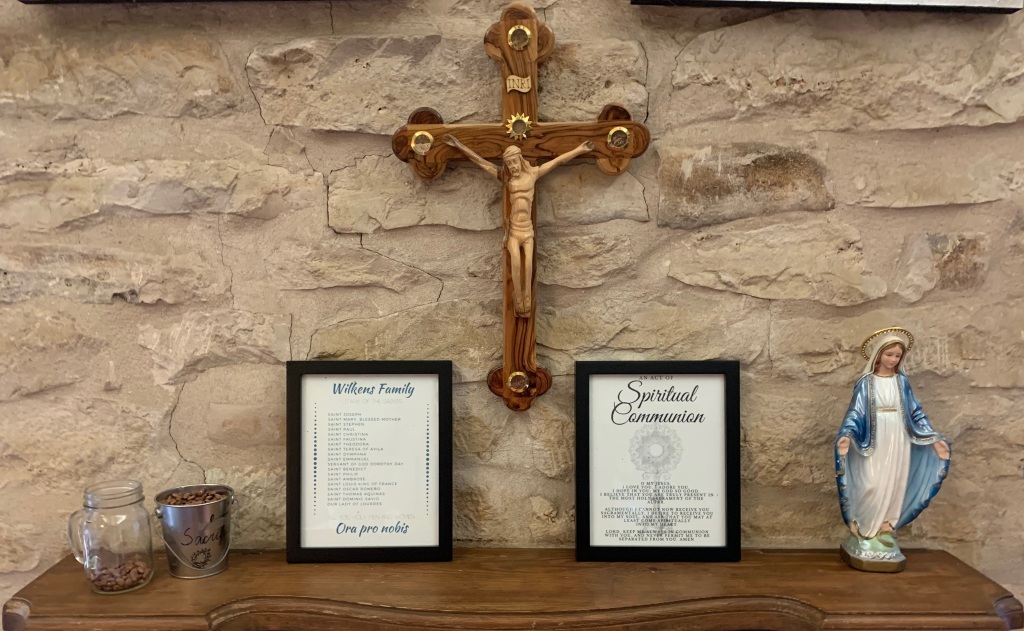 Crucifix hanging above mantel with Lenten sacrifice jar, family saints, spiritual communion prayer, and statue of the Blessed Mother.